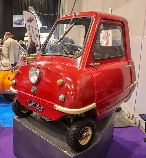 Tiny red car on a black plinth above a purple carpet, visitors behind. Opens a larger view