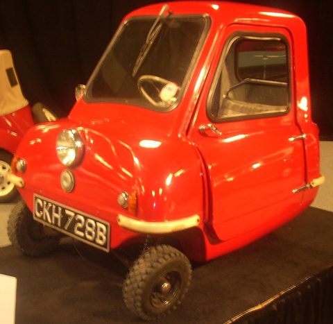 Tiny bright red car on a dark stand under show lights, seen from the front left. Opens a larger view