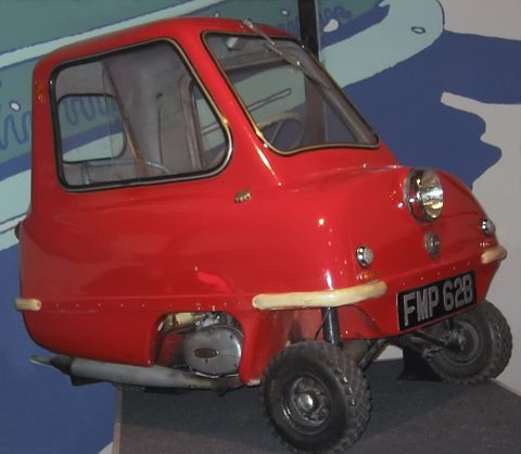 Tiny red car seen from the right side in a dim museum, the small engine showing under the sill. Opens a larger view