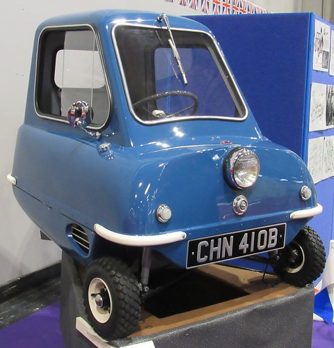 A tiny blue single-door microcar with one headlamp, two front wheels and chrome bumper strips on a low plinth. Opens a larger view