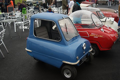Tiny blue car on wet tarmac next to a red domed Trident, café chairs behind. Opens a larger view
