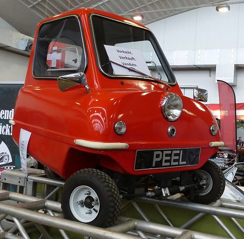 Tiny red car with a PEEL plate on a metal rack at a show, a notice in its window. Opens a larger view
