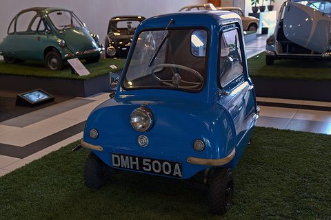 Tiny blue car on a strip of artificial grass, older microcars on display behind. Opens a larger view