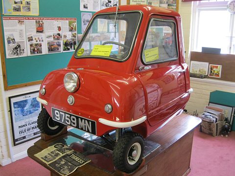 Tiny red car on a wooden stand in a small museum room with notice boards behind. Opens a larger view