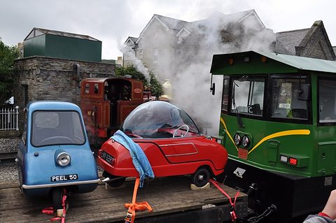 Tiny blue car and a domed red three-wheeler strapped to a flat wagon beside a green locomotive. Opens a larger view