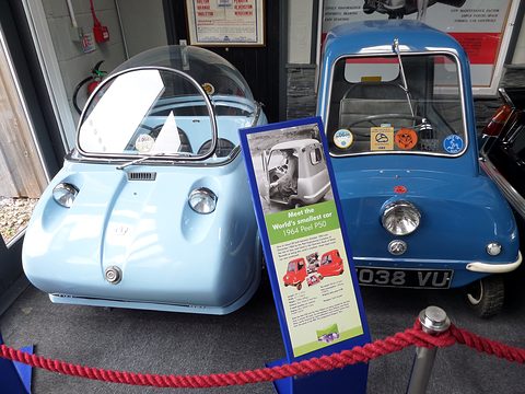Pale blue bubble car and a blue single-seat microcar behind a red rope, an information board between them. Opens a larger view