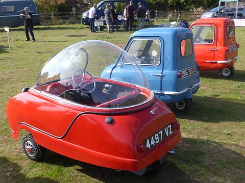 Red bubble car from the rear on grass, a blue and a red P50 parked beyond it. Opens a larger view