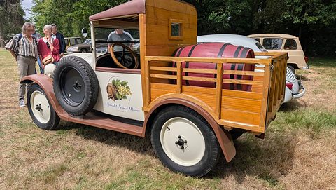 A cream and brown small truck with a varnished wooden cab and slatted bed holding a red barrel, a spare wheel on its flank. Opens a larger view