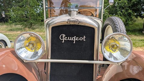 Close front view of a mesh radiator with Peugeot script between two large chrome headlamps with yellow lenses. Opens a larger view
