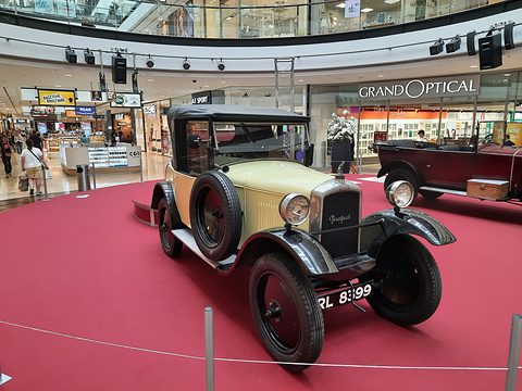 A small cream two-door car with black wings, a spare wheel on its side and a Peugeot radiator, inside a shopping mall. Opens a larger view