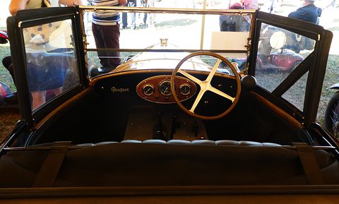 A four-spoke steering wheel before an oval wooden instrument panel with three round gauges, side screens open. Opens a larger view