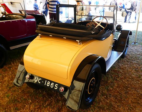 Rear three-quarter view of a small yellow cabriolet with a rounded tail, black wings and a folded black hood. Opens a larger view