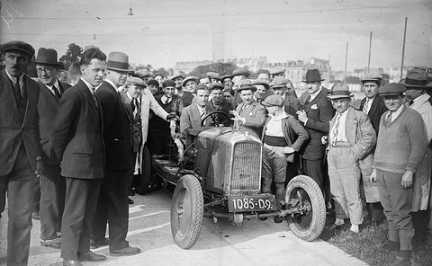A small open car with a slatted radiator and plate 1085-D9 surrounded by a dense crowd of men in caps and hats. Opens a larger view