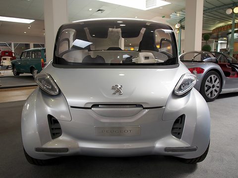 Front of a silver tall, short four-seat concept with slim lamps and glazed roof on a museum floor. Opens a larger view