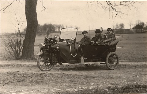 Three people in hats in a three-wheeled open tourer on a dirt road beside a bare tree. Opens a larger view