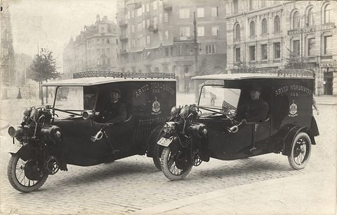 Two three-wheeled delivery vans with uniformed drivers parked side by side on a city street.