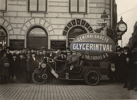 A three-wheeled van carrying a large oval soap sign, surrounded by a crowd of men and boys. Opens a larger view