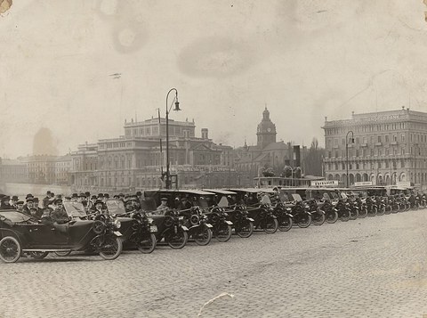 A long line of small three-wheeled cars with passengers on a cobbled quay, grand buildings behind. Opens a larger view