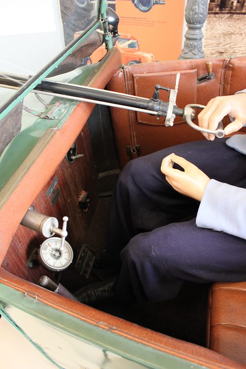 Looking down at a steering column, brown leather seats and a mannequin’s hands and legs. Opens a larger view