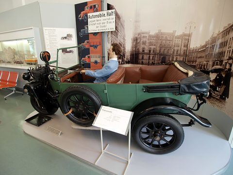Side view of a green open tourer with brown leather seats and a mannequin driver on a plinth. Opens a larger view