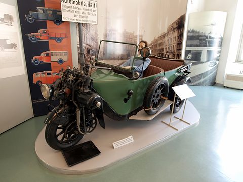 Green three-wheeled open tourer with a mannequin driver on a round plinth before period posters. Opens a larger view