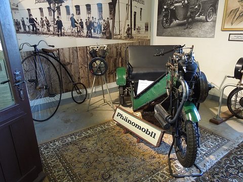 Green three-wheeled car on a patterned rug with a ‘Phänomobil’ sign and an old high-wheel bicycle. Opens a larger view
