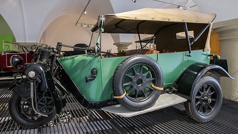 Green three-wheeled tourer with a tan hood and spare wheel on a metal grille floor under vaults. Opens a larger view