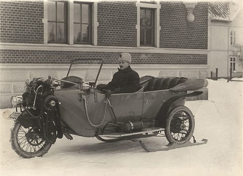 A man in a fur hat at the wheel of a three-wheeled open car standing on skis in snow. Opens a larger view