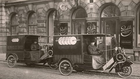 Two three-wheeled bread vans with drivers parked in front of a bakery with arched windows. Opens a larger view