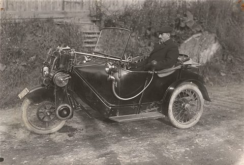 A man in a cap seated in a small three-wheeled open car, its engine over the front wheel. Opens a larger view