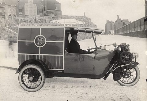 A three-wheeled van with a boxy body bearing a crown emblem, a uniformed driver at the wheel. Opens a larger view