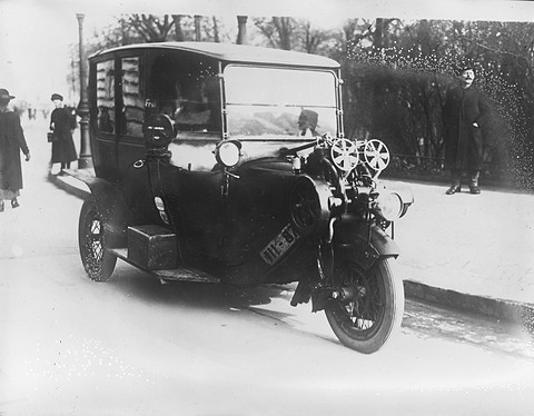 A boxy black three-wheeled cab with twin fans on the engine over its front wheel, a man on the pavement. Opens a larger view