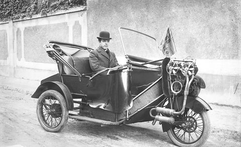 A man in a bowler hat in a three-wheeled open car with its engine over the front wheel, by a wall. Opens a larger view