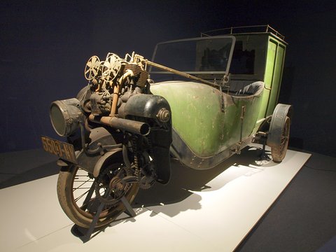 Front of a weathered three-wheeled van showing the engine and cooling fans above the front wheel. Opens a larger view