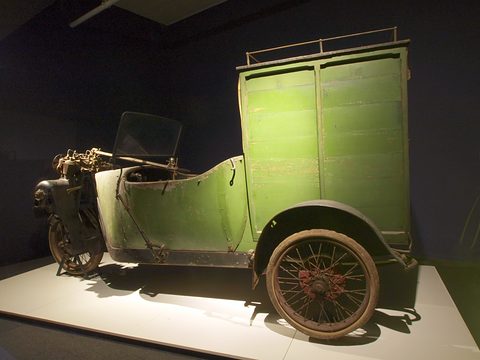Weathered green three-wheeled van with a tall box body on a white plinth in a dark room. Opens a larger view