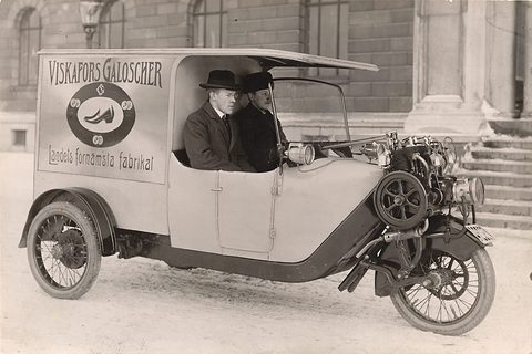 A three-wheeled van lettered ‘Viskafors Galoscher’ with two men in hats in front of a stone building. Opens a larger view