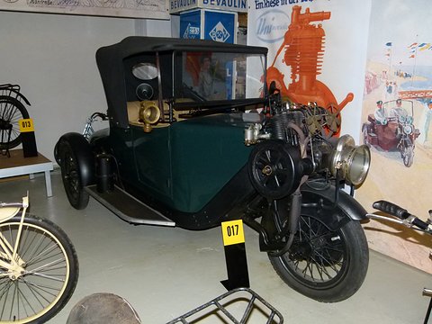 Dark green three-wheeled car with a black hood and its engine over the front wheel in a museum.