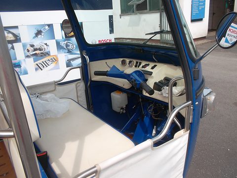 Handlebar steering, two round dials and toggle switches in a blue three-wheeler, a cream bench seat beside. Opens a larger view