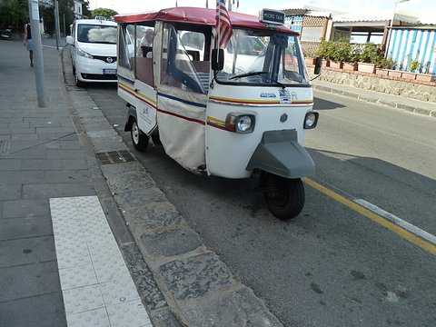 White and red striped three-wheeled open taxi with a canopy and flags parked at a kerb by a promenade. Opens a larger view