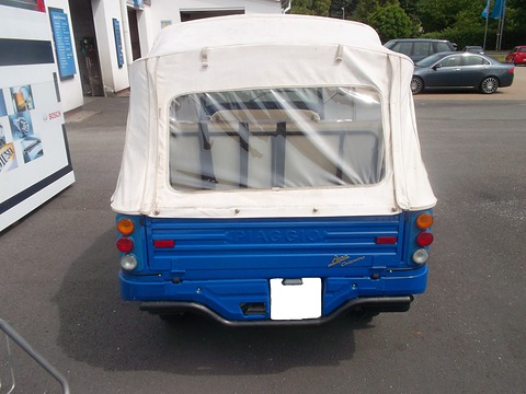 Rear of a blue three-wheeler with a white canvas hood and a blanked plate in a car park. Opens a larger view