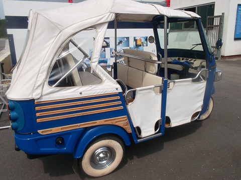 Side view of a blue three-wheeled open runabout with a white canvas canopy and wooden side strips. Opens a larger view