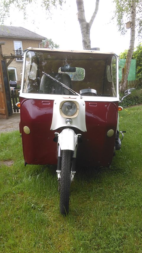 Head-on view of a maroon and cream three-wheeler with a single front wheel and headlamp, a white hood above, on grass.