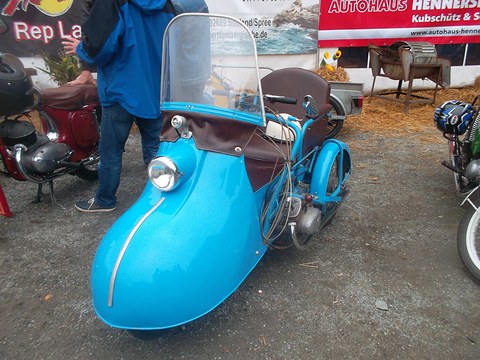 Bright blue single-seat invalid tricycle with a rounded nose, a tall windscreen and a brown leather seat.
