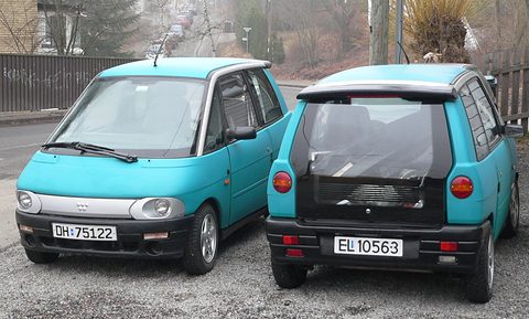 Two small turquoise two-door hatchbacks parked side by side on gravel, one facing the camera and one showing its tail. Opens a larger view