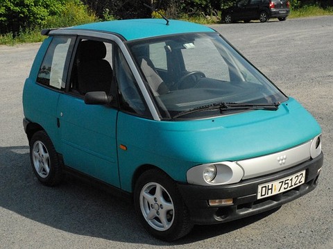 Turquoise small two-door car with a narrow grey grille band and black bumper on a gravel lot.