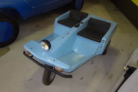 High view of a pale blue three-wheeled tub with two black seats flanking a central stick, a round headlamp on its nose. Opens a larger view