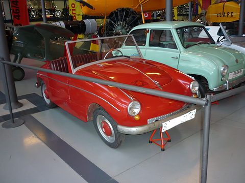Red open two-seater without doors, low windscreen and red seats, behind a steel rail beside a pale green Goggomobil.