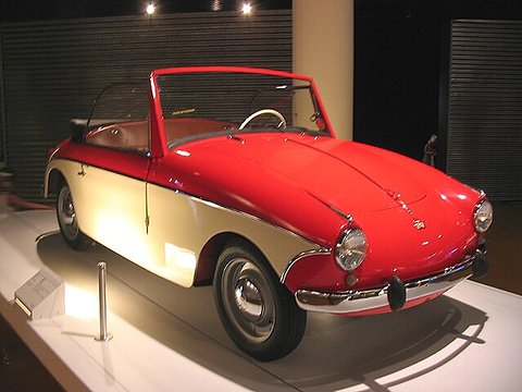 Red and cream open two-seater with chrome bumper and round headlamps, lit by spotlights against a dark slatted wall. Opens a larger view