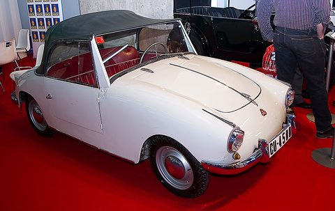 White open two-seater with a black folded hood and red seats, on red carpet next to a black vintage tourer. Opens a larger view