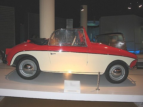 Side view of a small red open car with a cream lower body and chrome wheel discs on a white plinth.